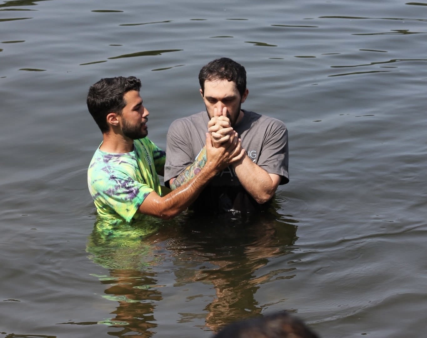 Zachary Denning being baptized in open water, hands clasped in prayer, held steady by a friend